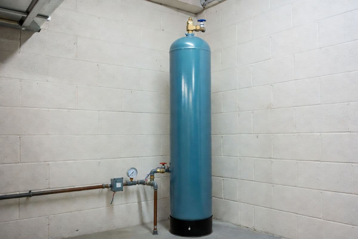 Blue well water pressure tank installed in a residential basement utility room with copper pipe connection, pressure gauge, and pressure switch visible.